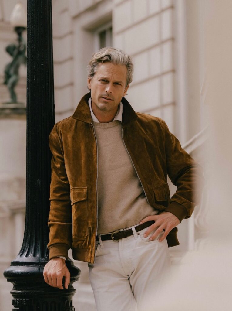 Giacca bomber in renna uomo colore tabacco indossata con maglione beige e pantaloni bianchi.